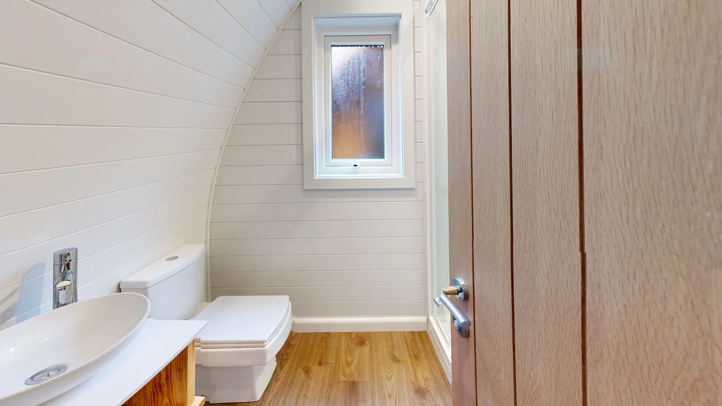 Small bathroom with wood flooring, a white toilet, a small white sink, a window, and wood-paneled wall.