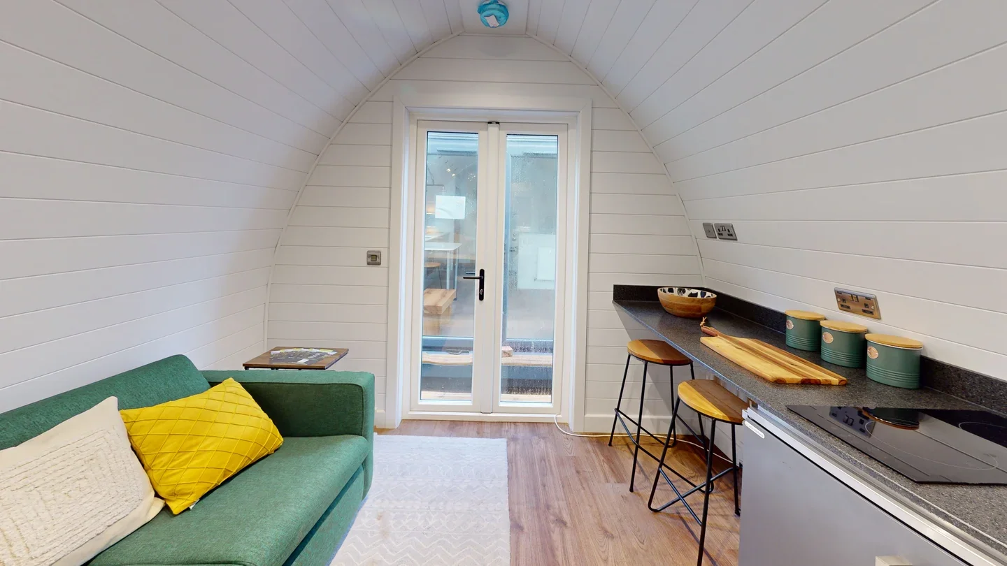 Small apartment kitchenette with green sofa, yellow cushion, white pillows, wooden stools at a black countertop, and a sliding glass door leading outside.