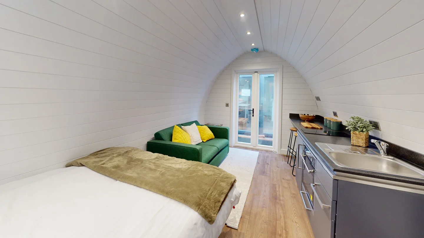 Small, cozy studio apartment with a curved ceiling, featuring a bed with a blanket on it, a green sofa with yellow and white pillows, a compact kitchen with black countertops, and sliding glass doors leading outside.