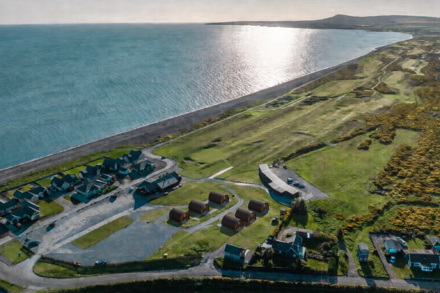 Aerial view of a coastal area with houses, green fields, and a shoreline adjacent to a large lake or sea, with hills in the distance and sunlight reflecting off the water.
