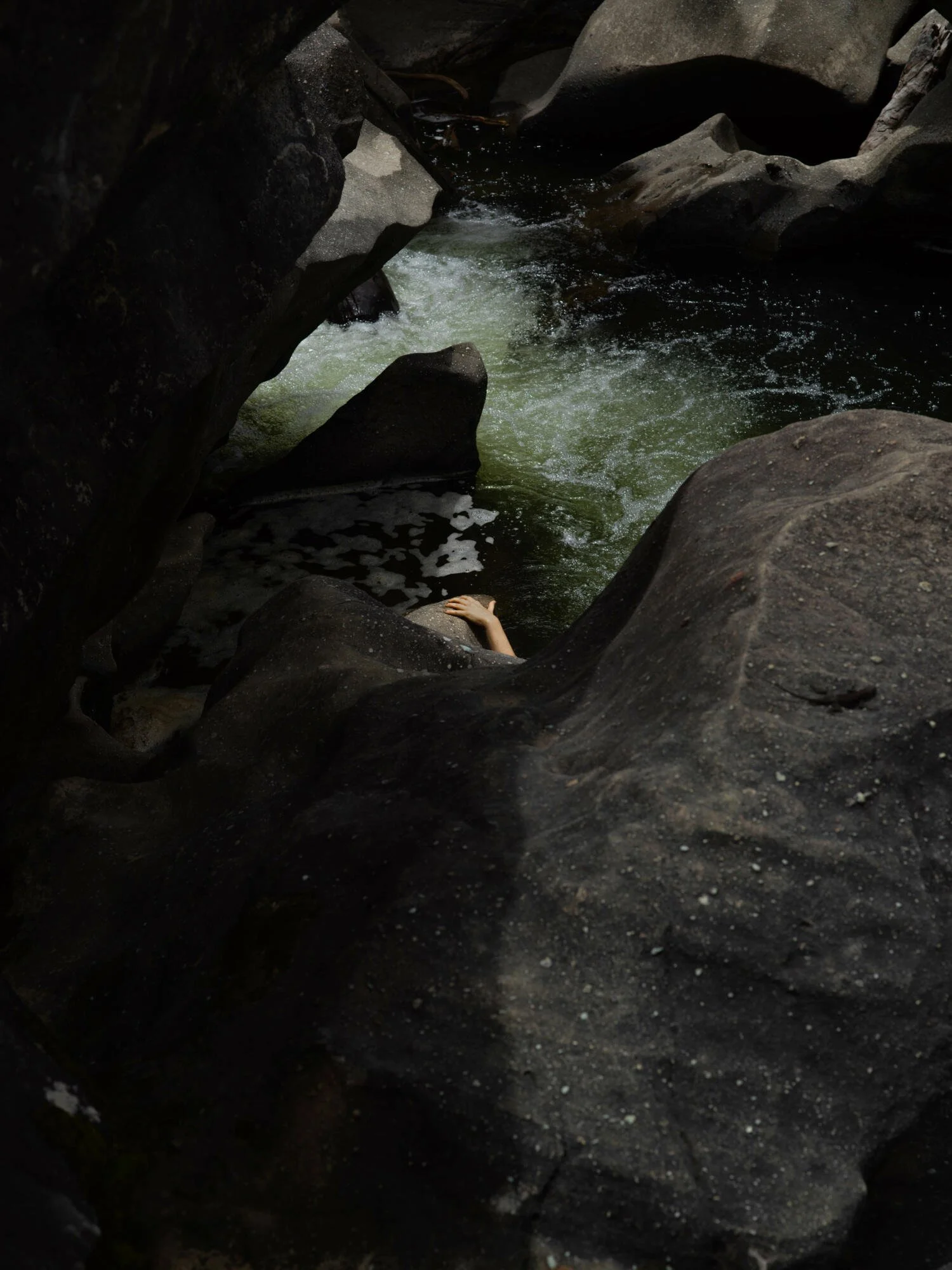 A person is reaching into a small, rocky canyon with flowing water, surrounded by large dark rocks and boulders.