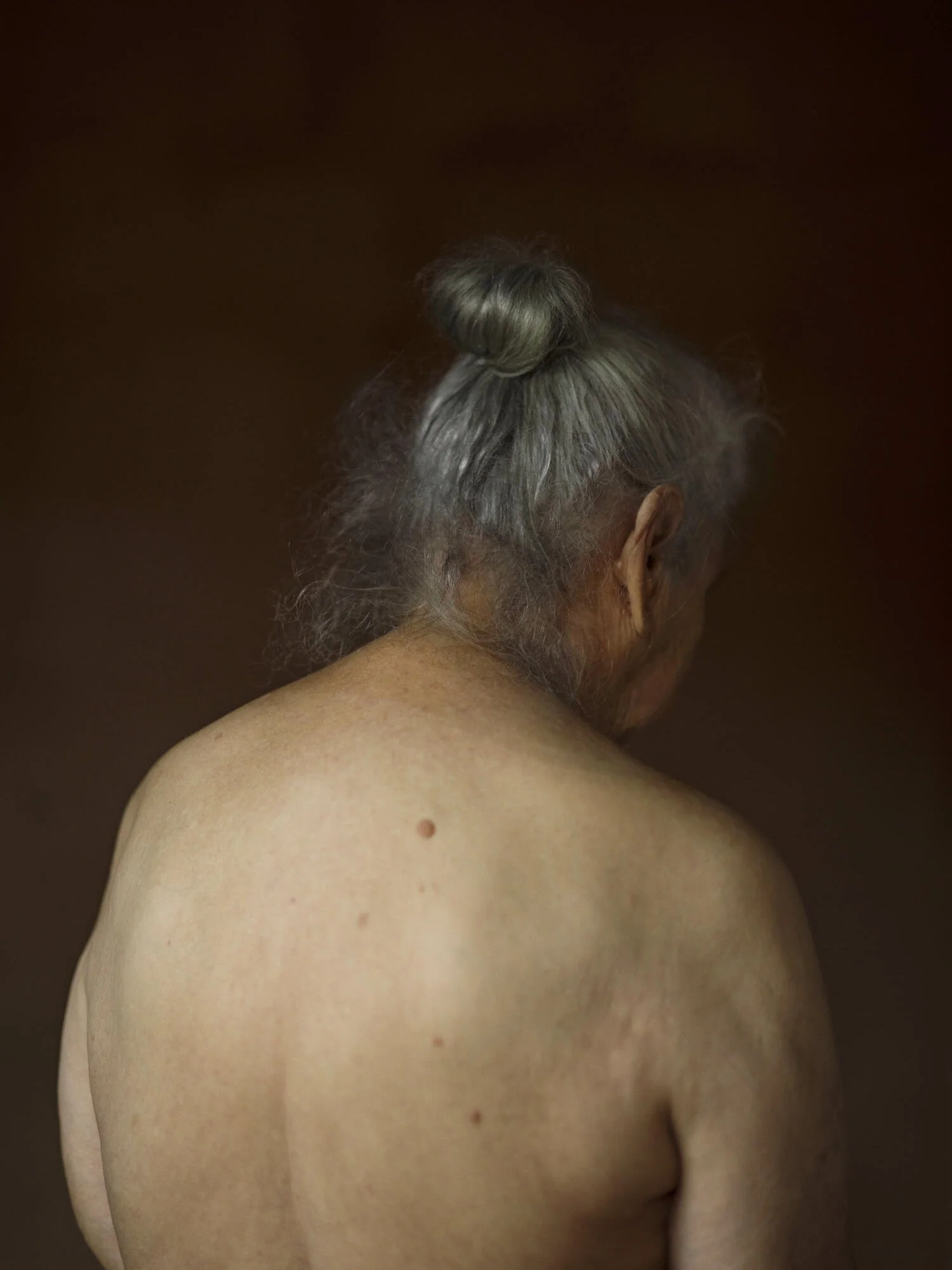 Back view of an elderly woman with gray hair tied in a bun, showing her shoulder, neck, and partial head against a dark brown background.