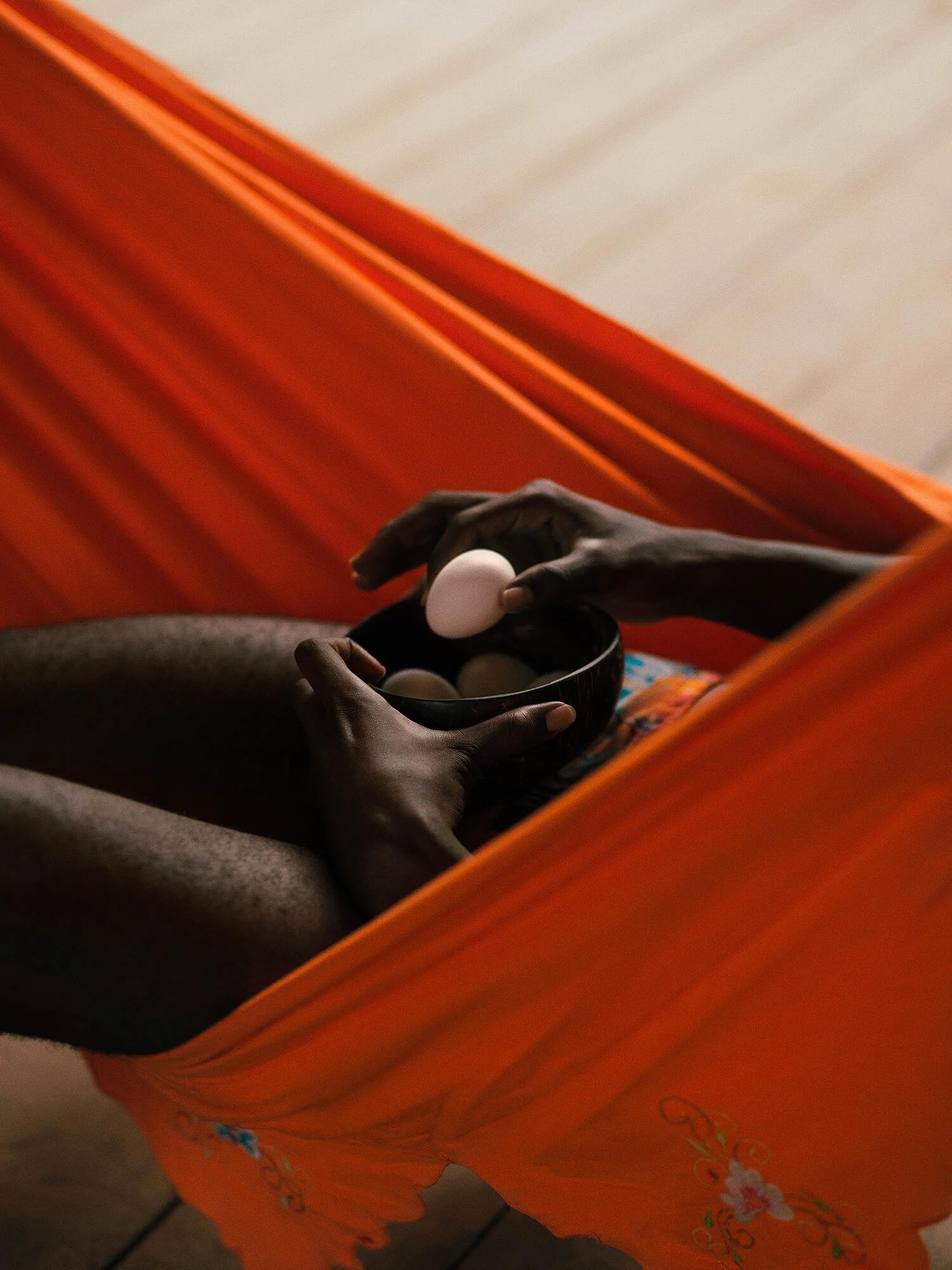 A person's hand holding a bowl with eggs above their lap, seen from above, with an orange chair or couch in the background.