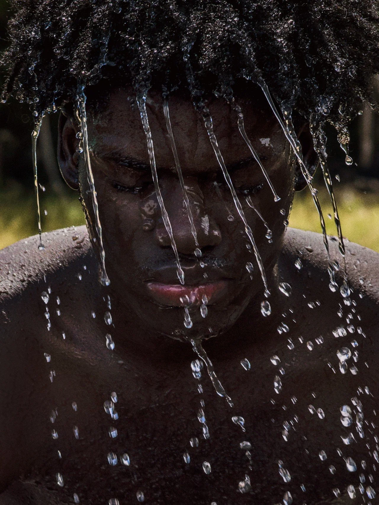 A person with dark skin and short, curly hair taking a shower. Water is cascading down their face and shoulders with their eyes closed.