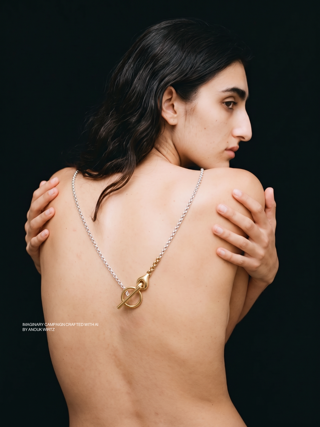 A woman with dark, wavy hair, topless, gazing to her right, holding her shoulders with her hands, wearing a necklace with a gold and silver toggle clasp and a small diamond charm, against a black background.