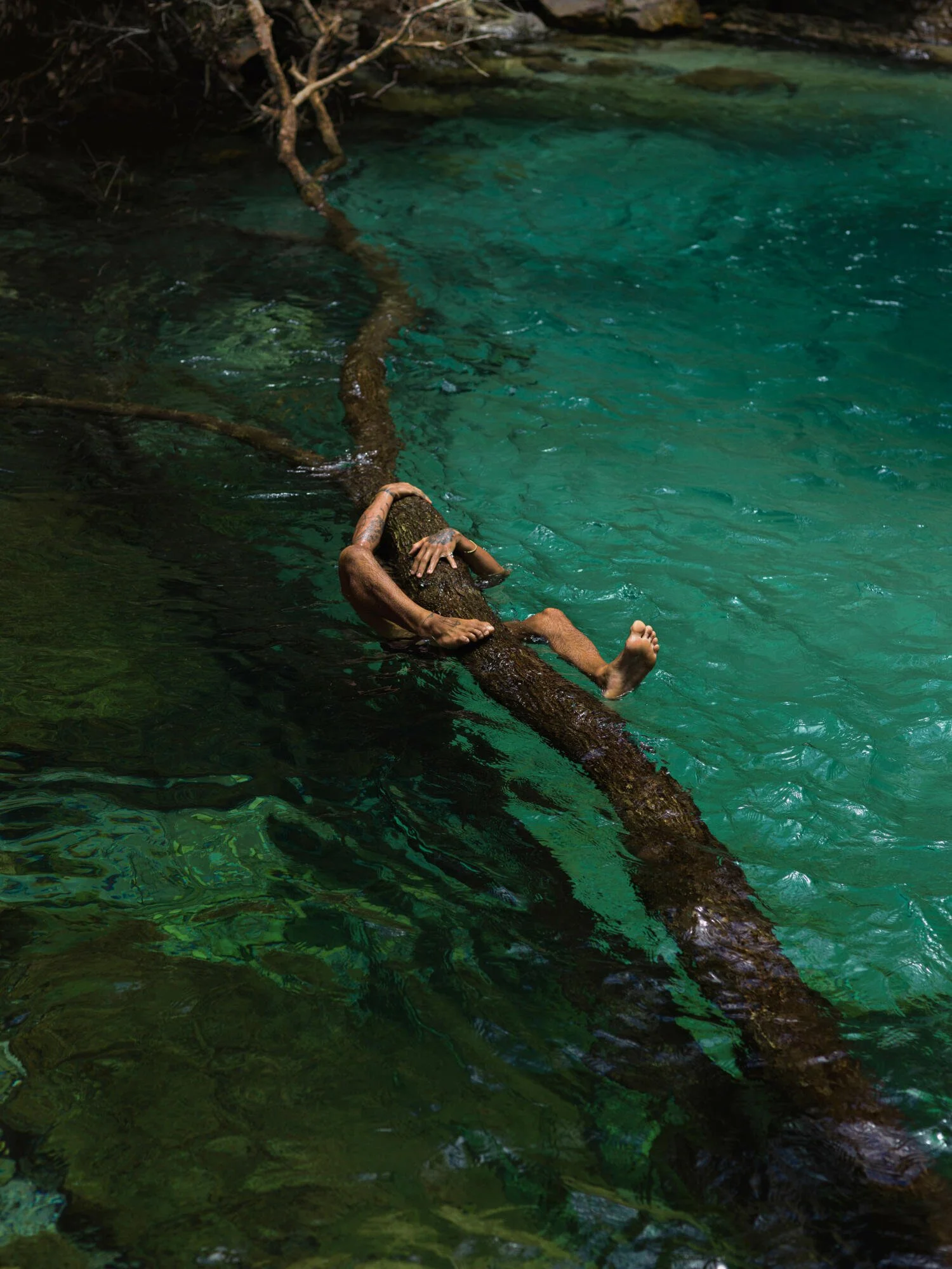 Person with tattoos lying on a fallen tree trunk by a clear, turquoise stream in a natural setting.