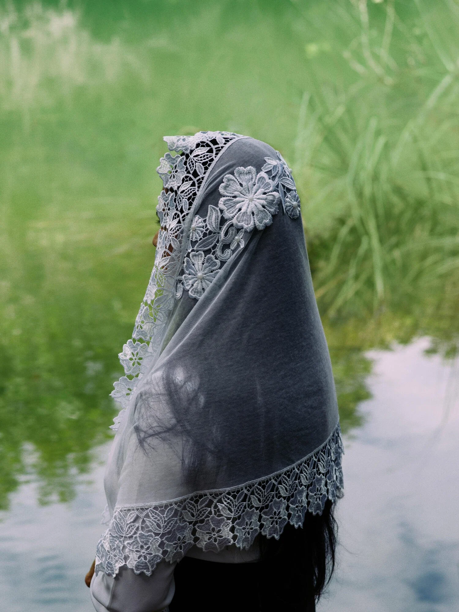 A person wearing a lace shawl and a gray head covering stands near a body of water with green foliage in the background.