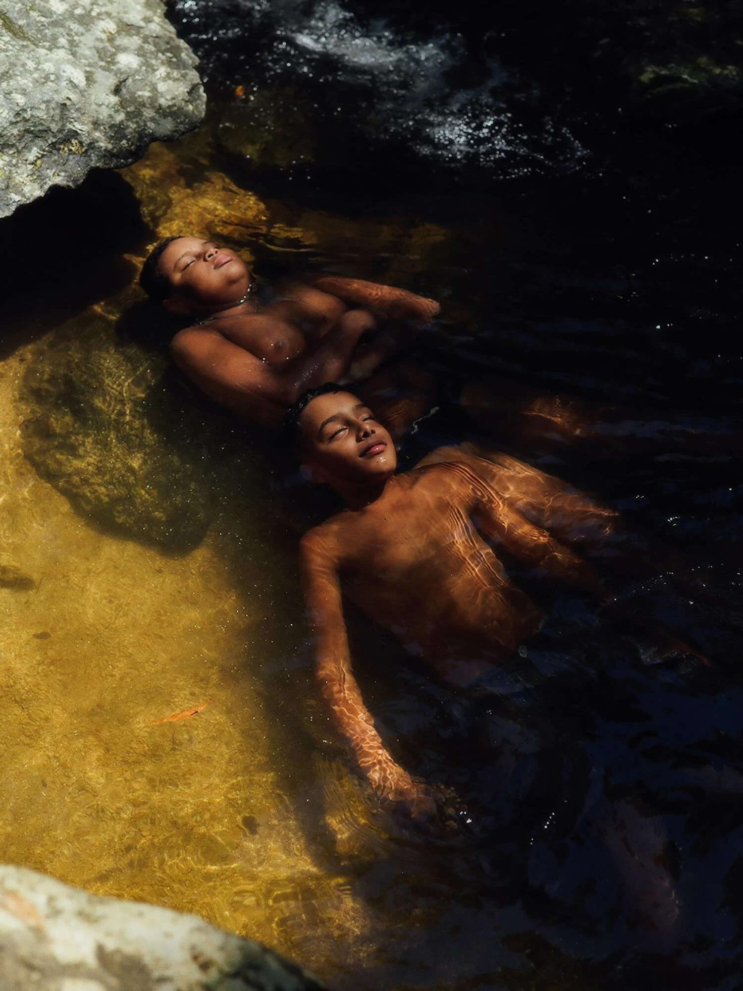 Two individuals are submerged in water, relaxing with their eyes closed, near rocks with moss and water bubbles visible.