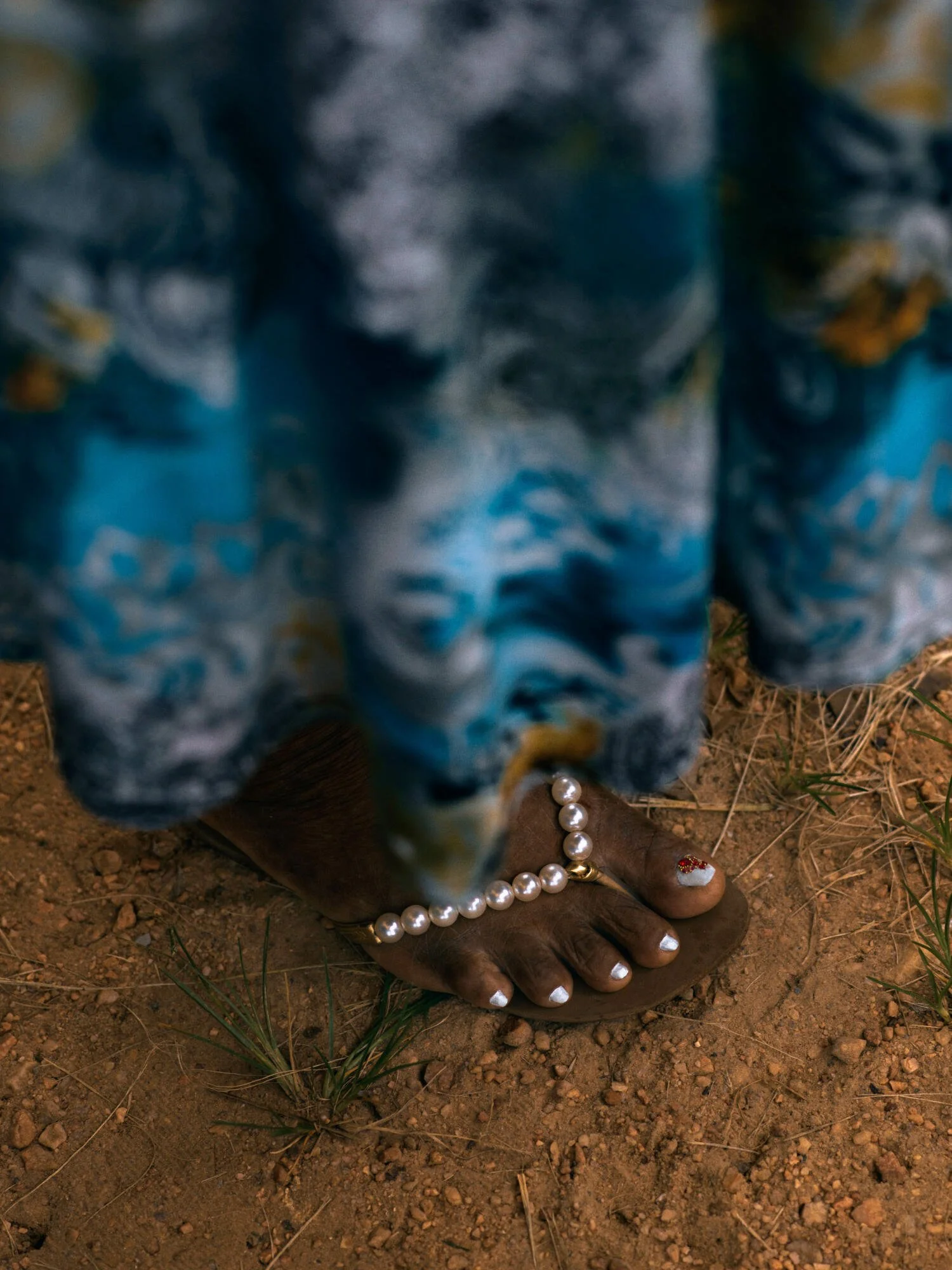 A person wearing a blue patterned dress with gold accents standing on dirt ground, with one foot visible wearing brown sandals and white painted toenails, surrounded by a string of pearl necklaces, small red and white decorative shoes, and some green