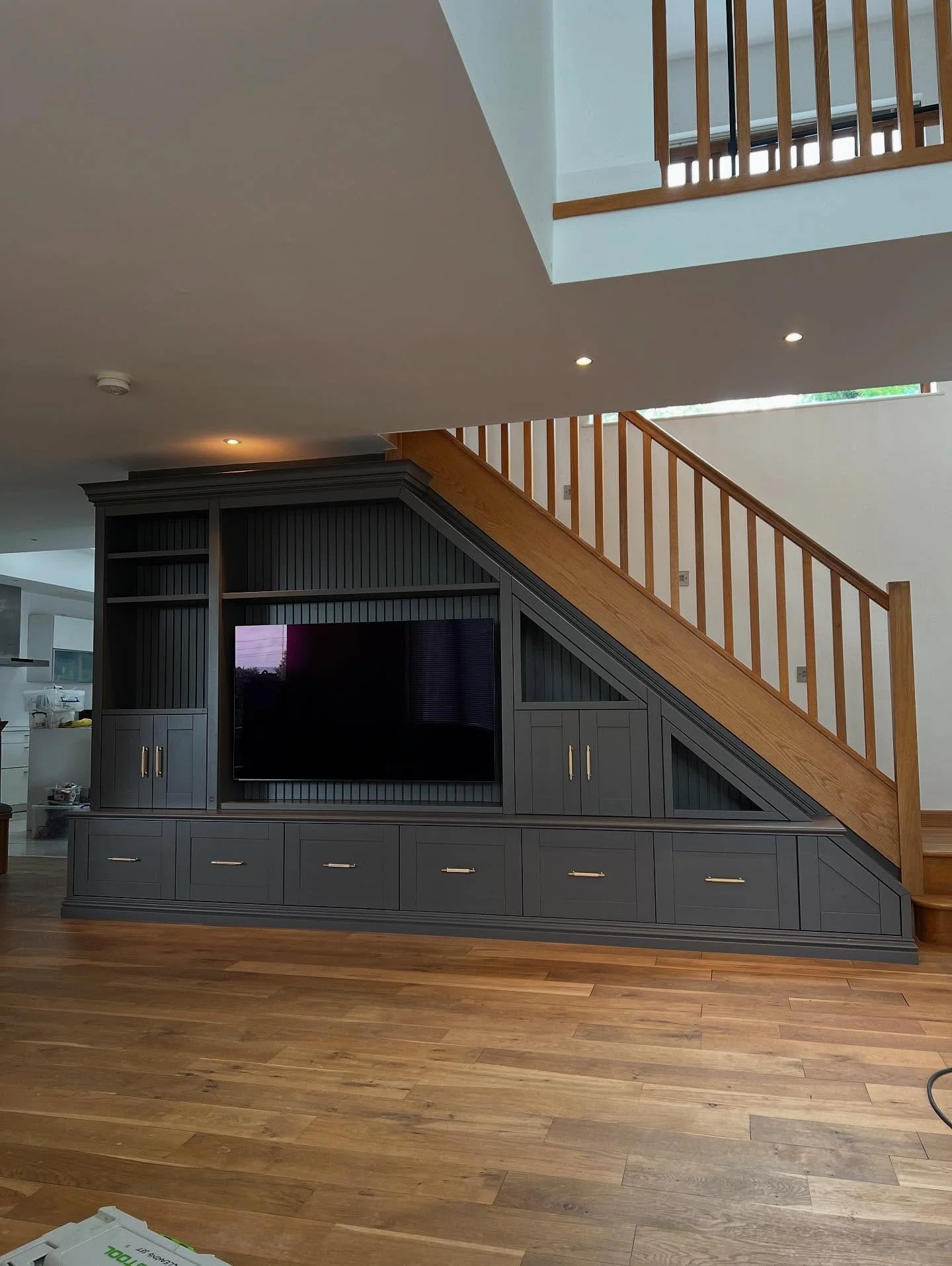 A modern living room with a custom built-in entertainment center painted dark gray, featuring a flat-screen TV and several storage cabinets, under a staircase with wooden railings on the second floor.