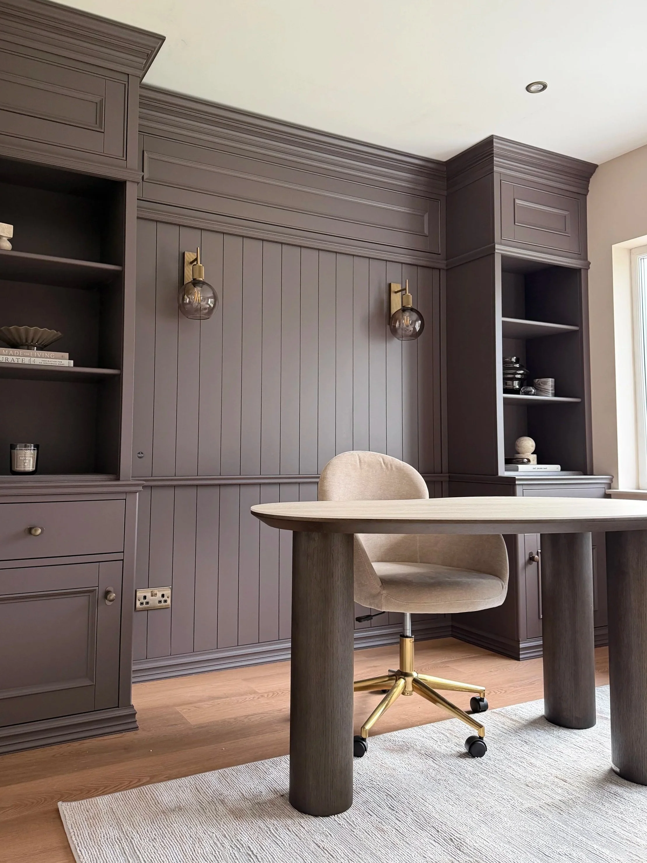 A modern home office with a round wooden table, beige swivel chair, gray paneling wall, wall-mounted lights, built-in shelves with decorative items, and a window letting in natural light.