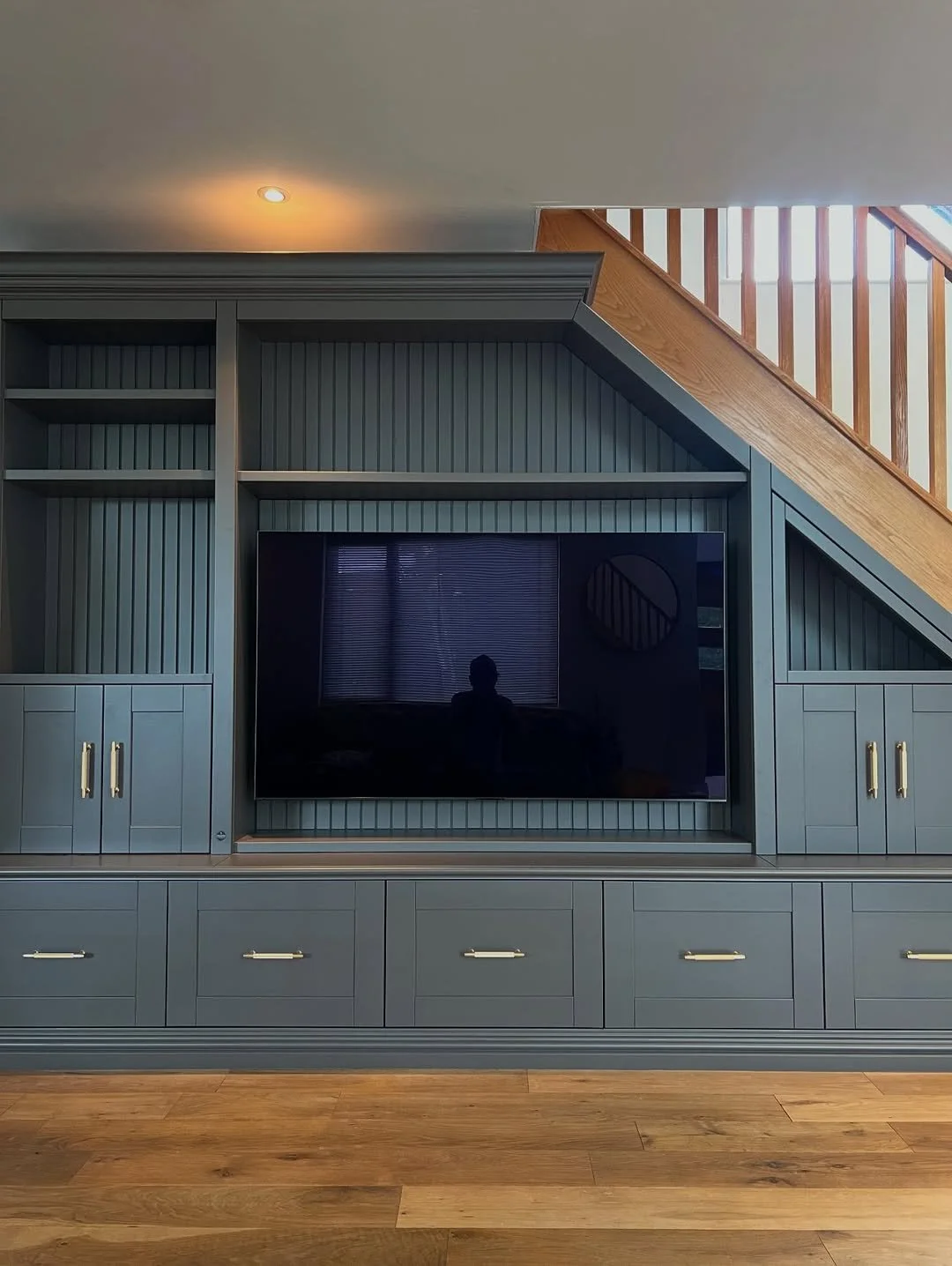 Built-in blue cabinet with open shelves, closed cabinets with gold handles, and a mounted flat-screen TV, in a room with wooden flooring and stairs with wooden railing in background.