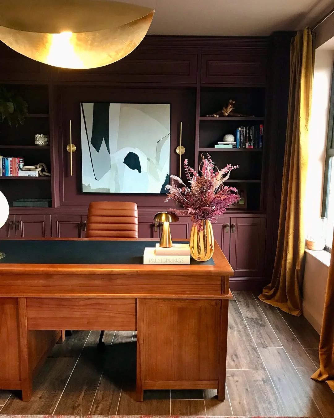 A home office with a wooden desk, a brown leather chair, a dark purple bookcase with books and decorations, a large colorful painting of a horse, a gold vase with pink flowers, and a window with yellow curtains.
