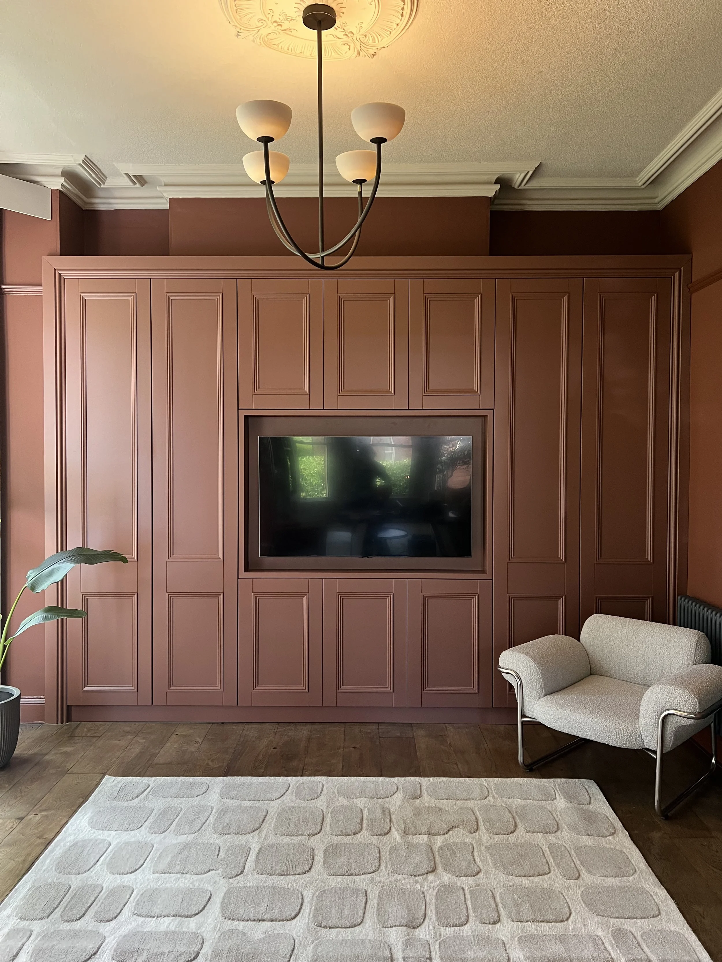 Living room with a large built-in wall unit painted in a muted pink color, housing a flat-screen TV in the center. There is a modern chandelier with five frosted glass shades hanging from the ceiling. A white armchair with black metal legs sits to the right, and a potted plant with large green leaves is on the left. A light-colored rug with a geometric pattern covers part of the wooden floor.