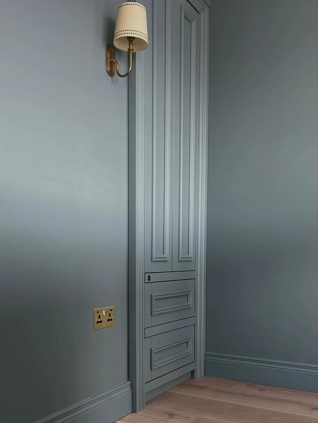 A corner of a room with a light blue wall, a beige wall-mounted lamp, a closed white wooden cabinet with detailed molding, a beige electrical outlet, and wooden flooring.