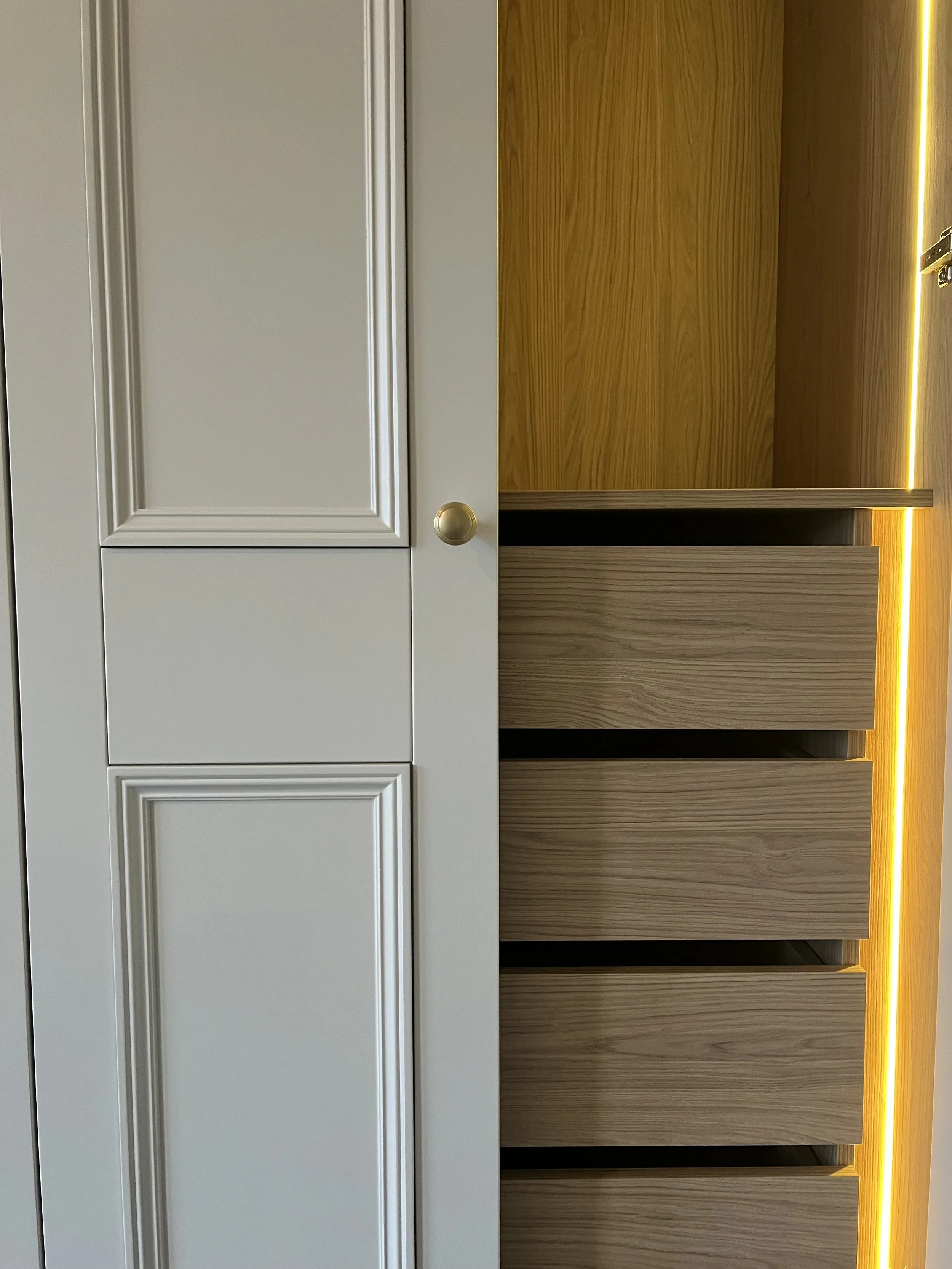 Close-up of a modern built-in wooden wardrobe with four open drawers, light wood finish, and a section with a light-colored cabinet with decorative molding on the panels and a round gold knob.