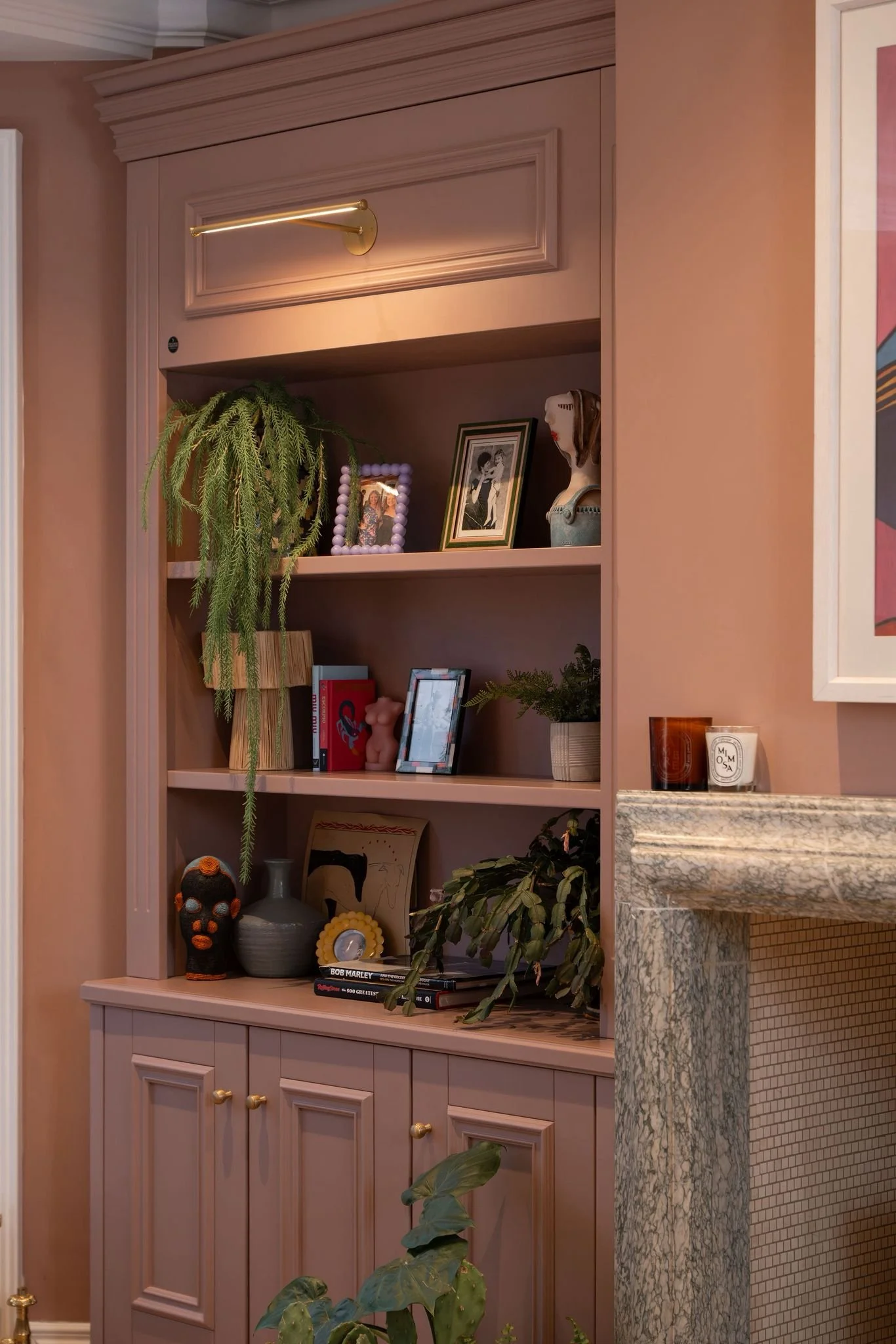 A pink built-in bookshelf with decorative items, books, framed photos, plants, and a gold reading light, adjacent to a marble countertop.