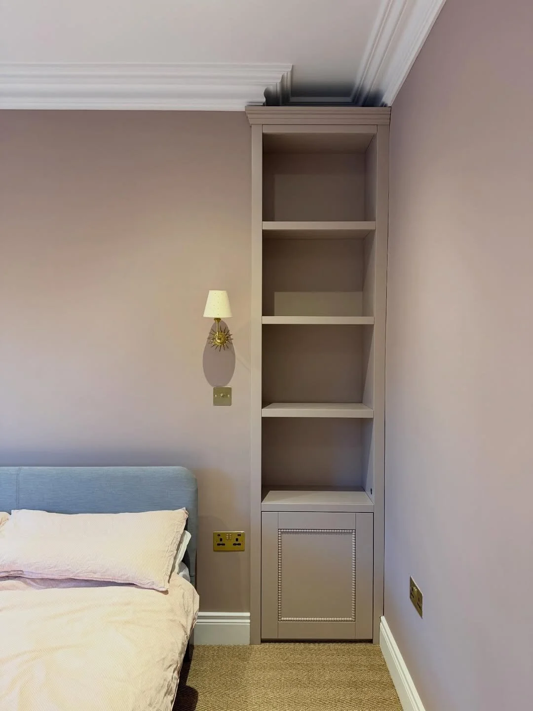 Empty built-in bookshelf with four shelves and a cabinet at the bottom, located next to a bed with a light blue headboard, in a room with beige walls and crown molding, with electrical outlets and a wall-mounted lamp nearby.