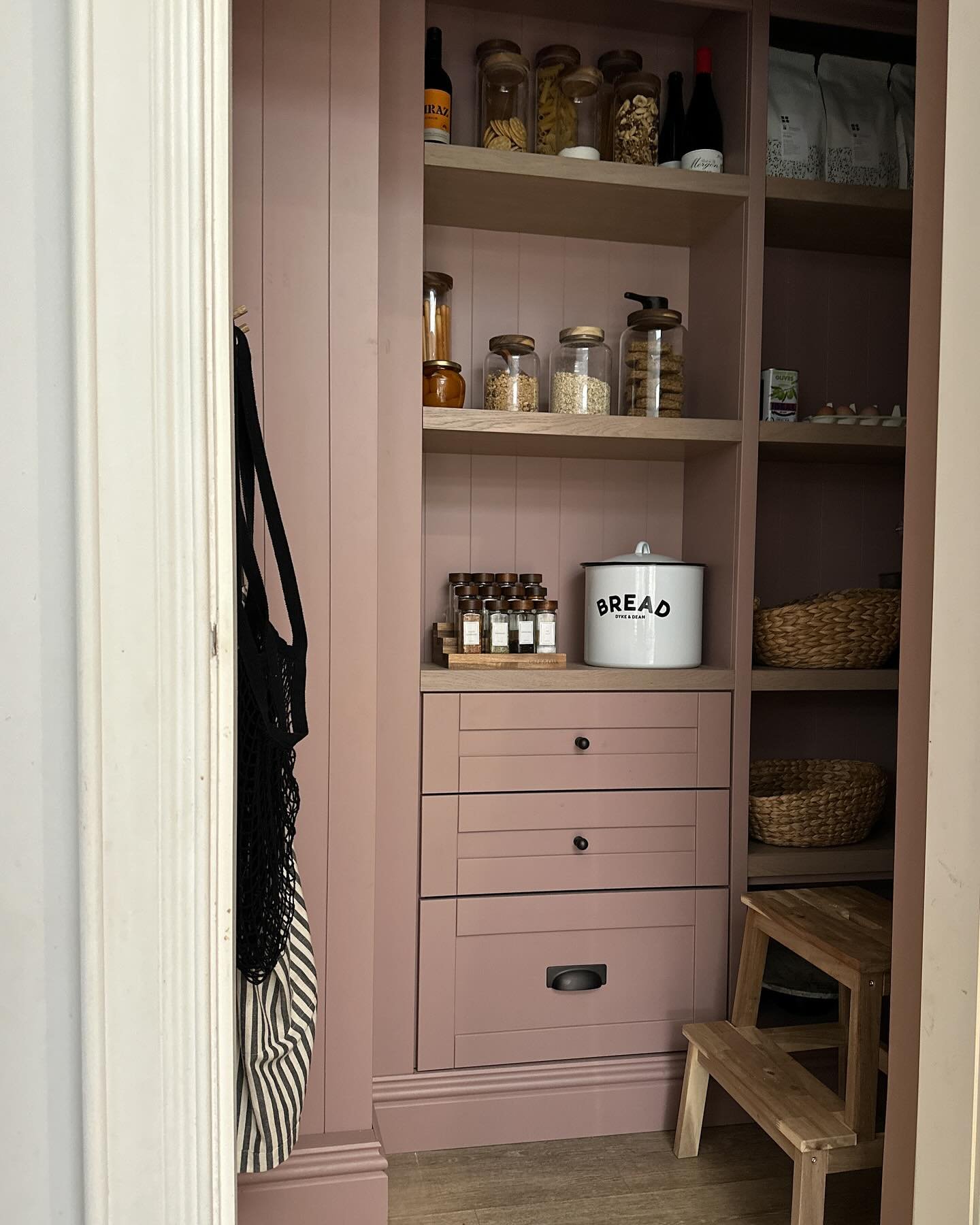 A pantry with pink cabinetry filled with jars of pasta, grains, and spices, a bread container, wicker baskets, a small wooden step stool, and eggs in the background.