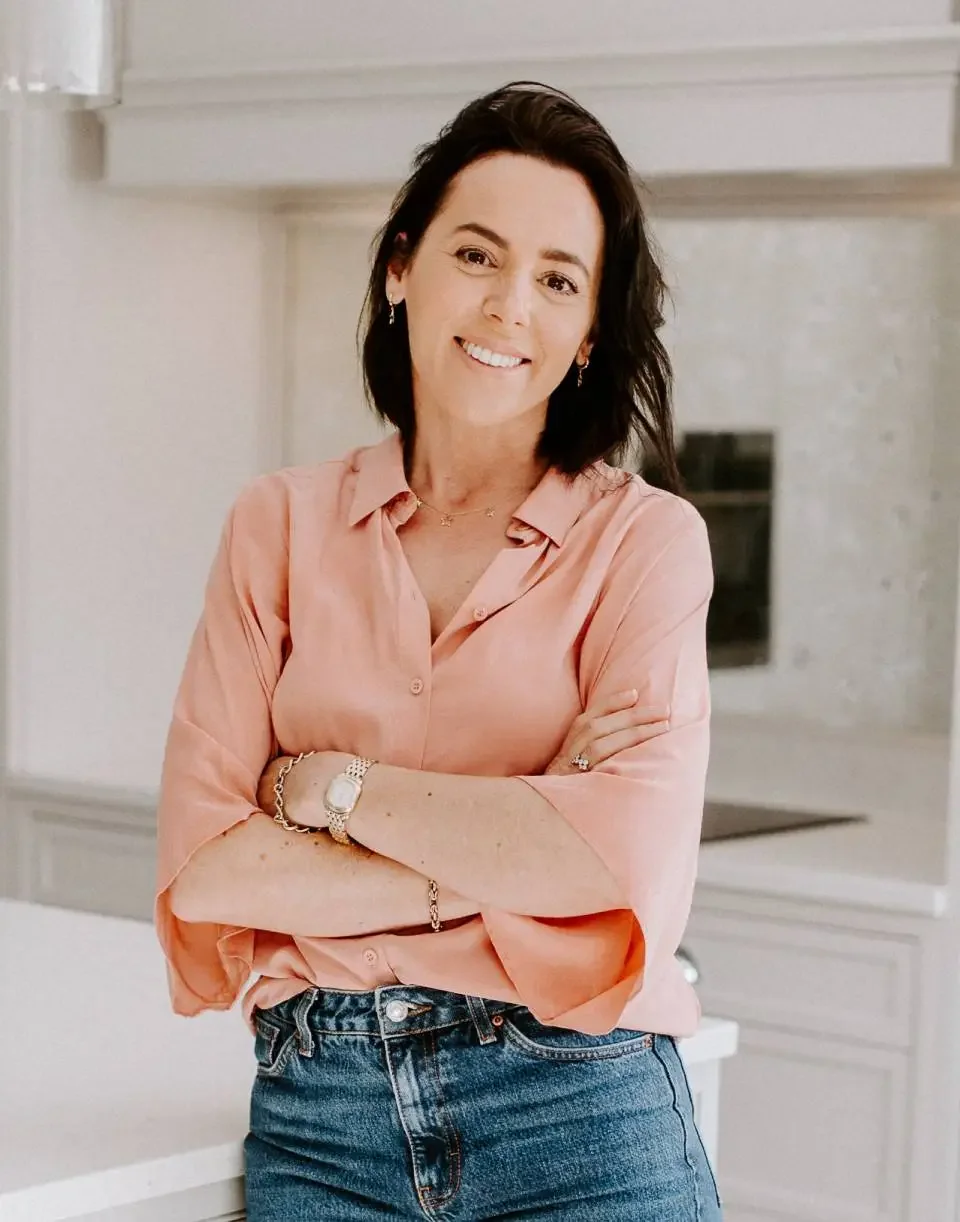 A woman with dark hair, wearing a peach-colored blouse and blue jeans, smiling with her arms crossed, standing in a bright kitchen.