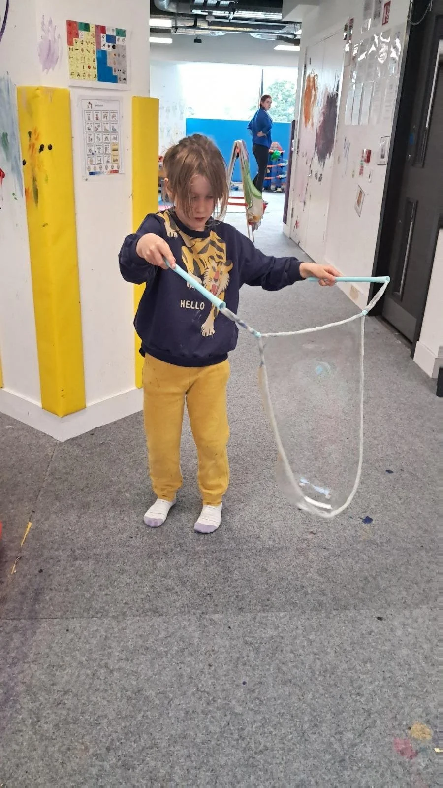 A young girl with shoulder-length hair wearing a dark blue sweatshirt with a tiger graphic and yellow pants, playing with a large bubble wand in an indoor classroom or art room.