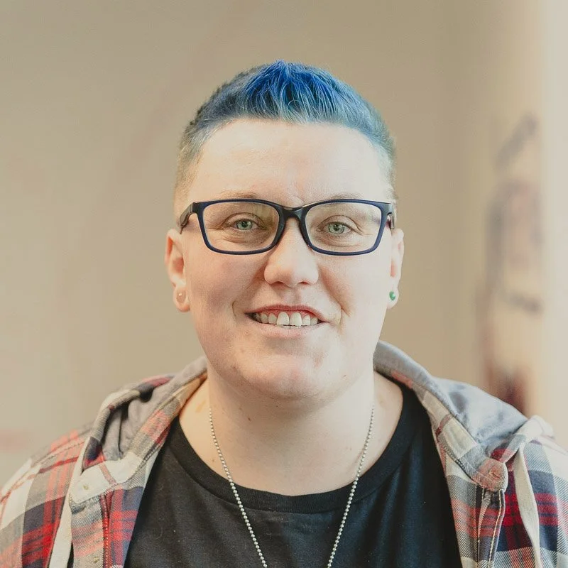A person with short hair dyed blue, wearing glasses, a black t-shirt, and a plaid hoodie, smiling for the camera.