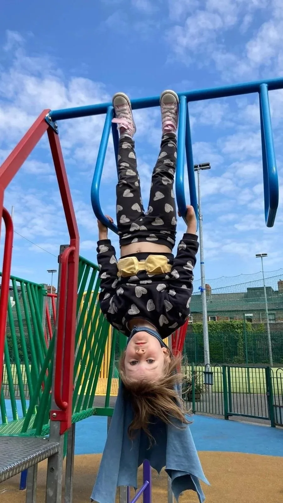 A young girl hanging upside down on playground bars with a blue sky and few clouds in background.
