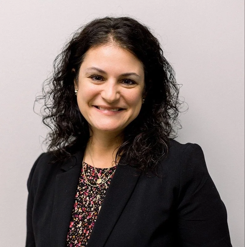 A woman with dark, curly hair smiling at the camera, wearing a black blazer and a floral top, standing against a plain light-colored background.