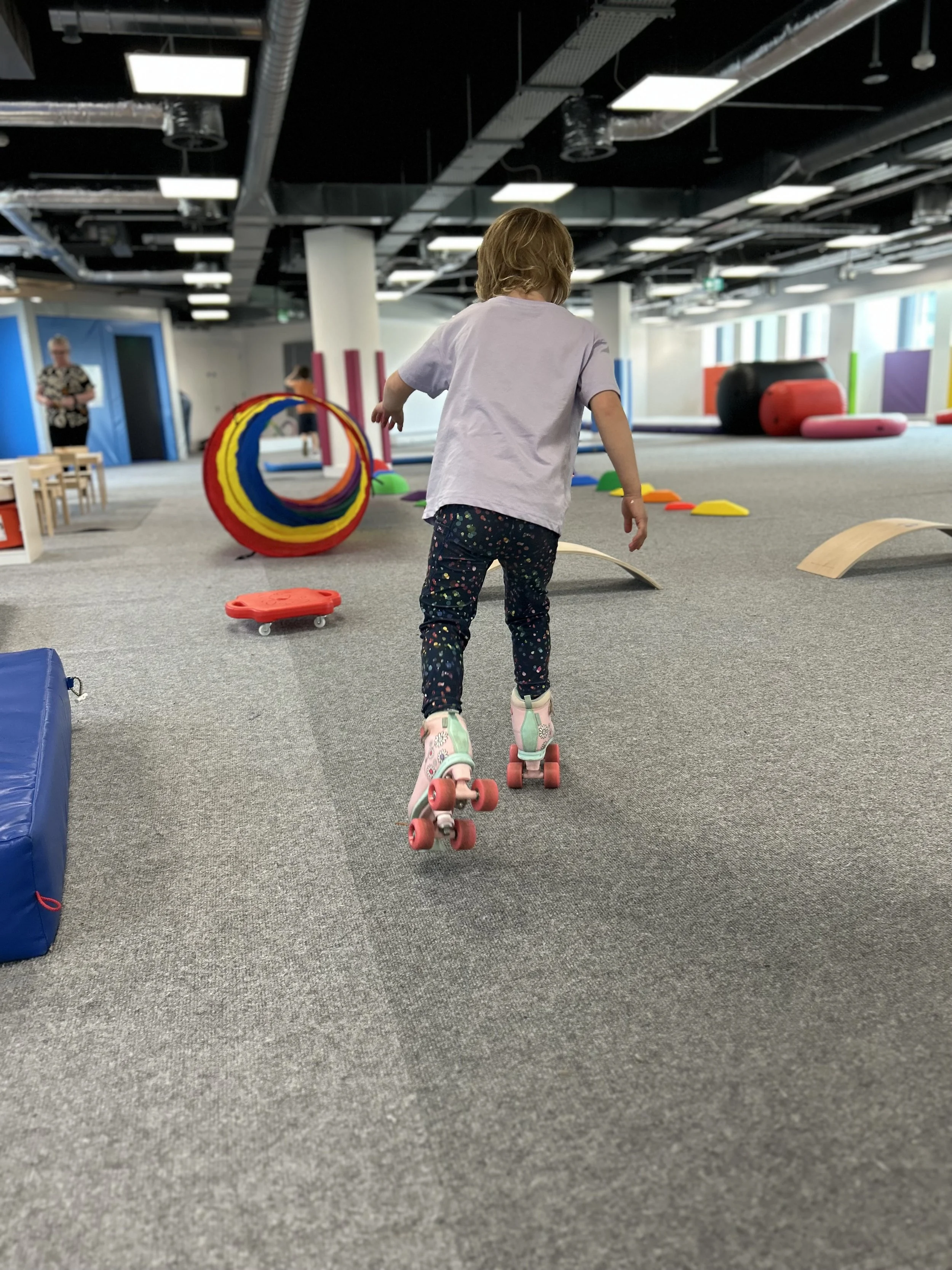 A young girl rollerskating through an indoor play area with various play equipment such as ramps, tunnels, and foam blocks.