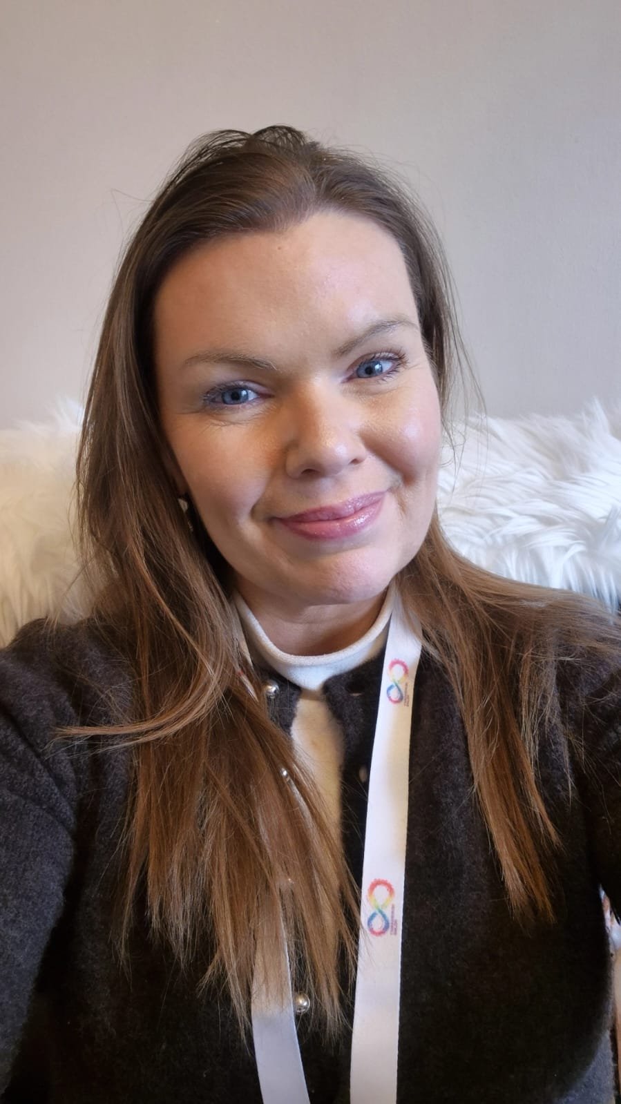 A woman with long brown hair and blue eyes smiling for a selfie. She's wearing a dark jacket with a white collar and a lanyard with colorful infinity symbols around her neck. Behind her is a fluffy white pillow and a plain beige wall.
