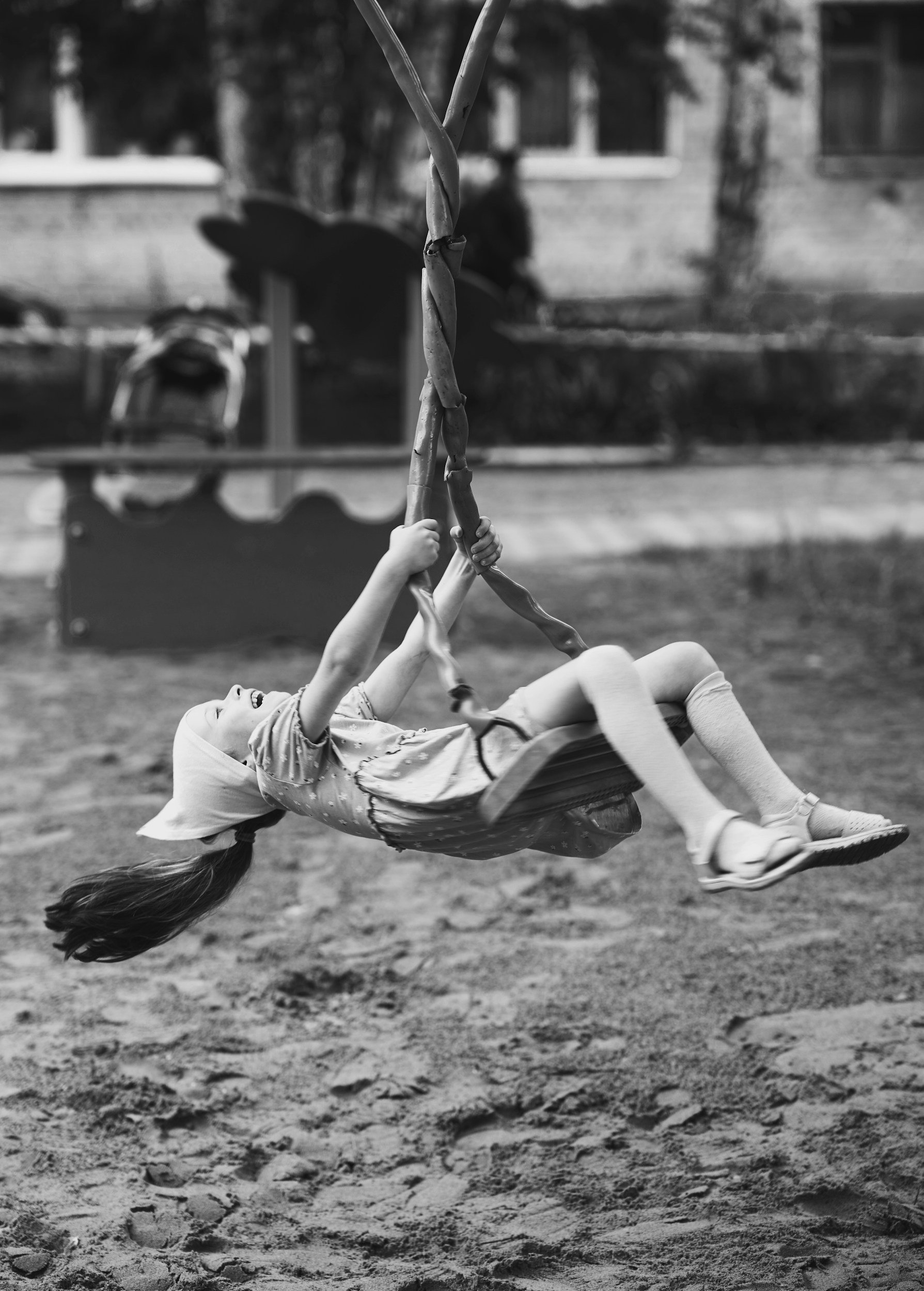 A young girl wearing a headscarf playing on a swing in a playground, captured in black and white.