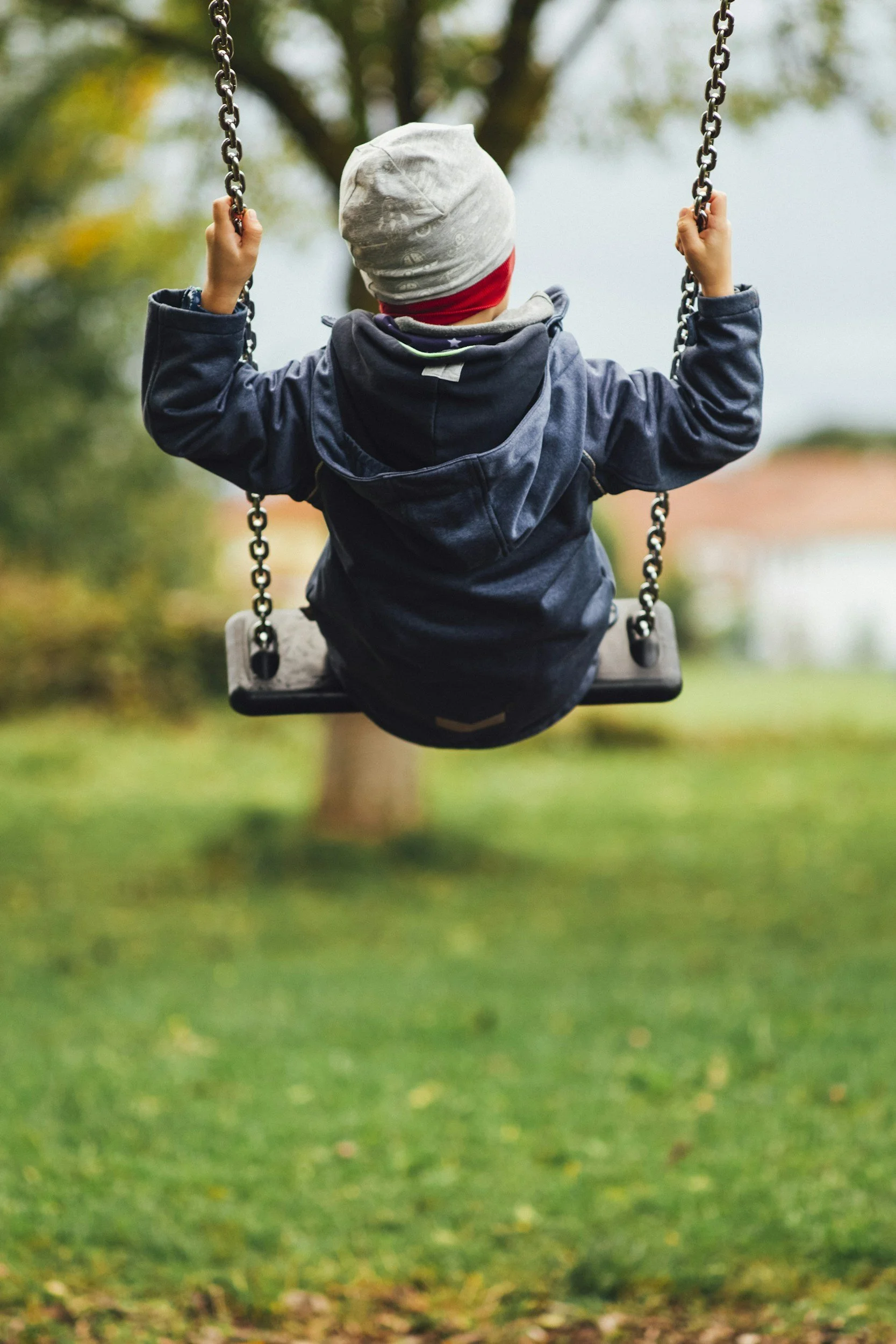 Child in a gray beanie and dark jacket sitting on a swing in a park with green grass and trees in the background.