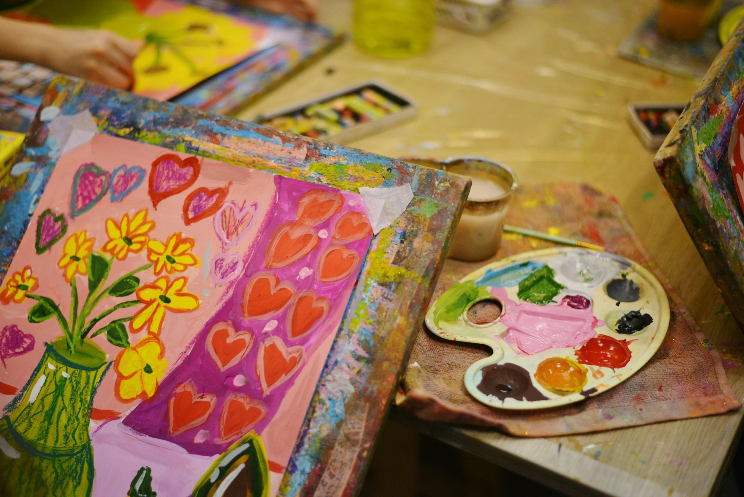 Painted canvas with a yellow vase containing yellow flowers and hearts on a pink background, surrounded by painting supplies including a palette with assorted colors and a cup of water.