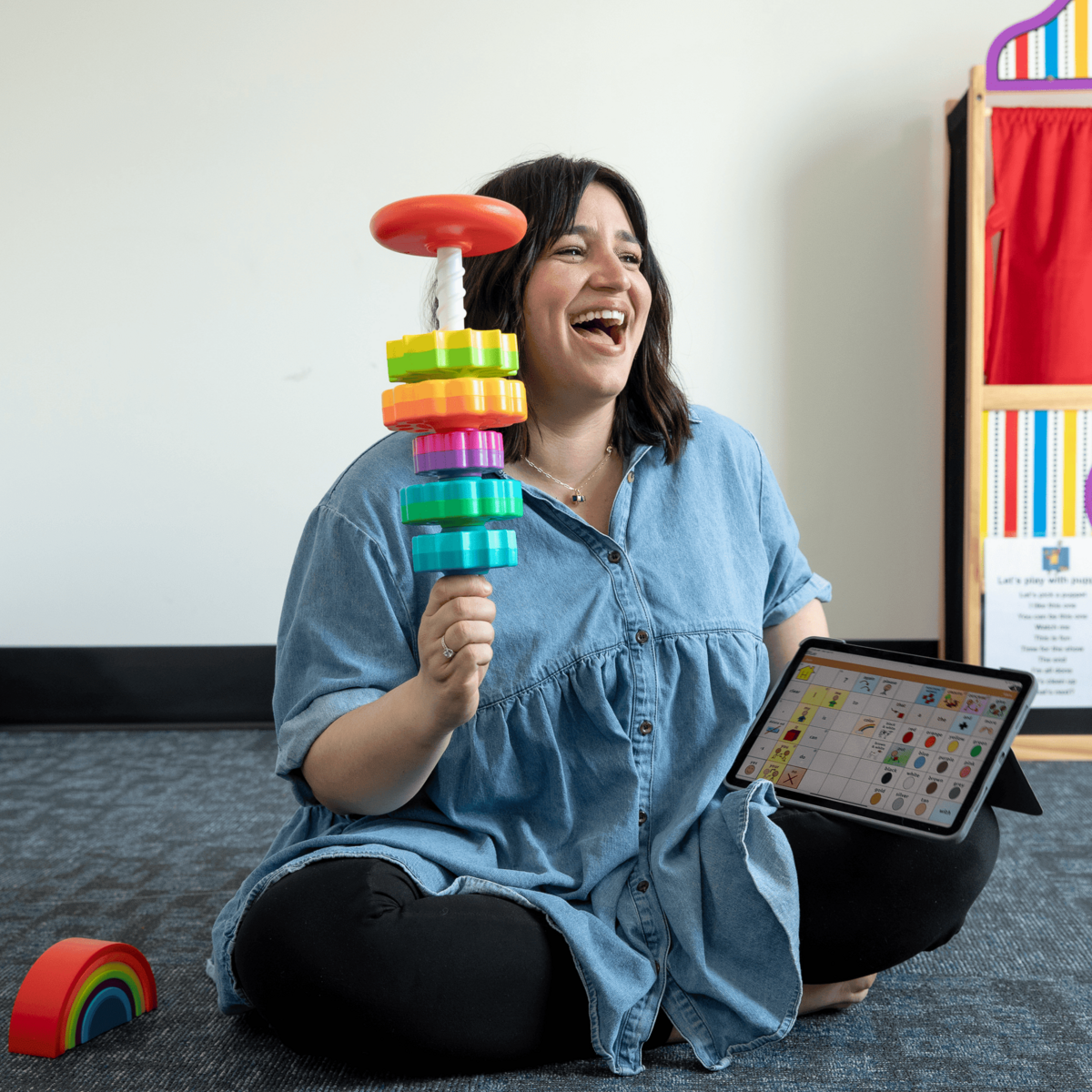 Woman sitting cross-legged on the floor, holding a colorful stacking ring toy in one hand and a tablet with a educational game screen in the other, laughing and enjoying her activity in a classroom or playroom.