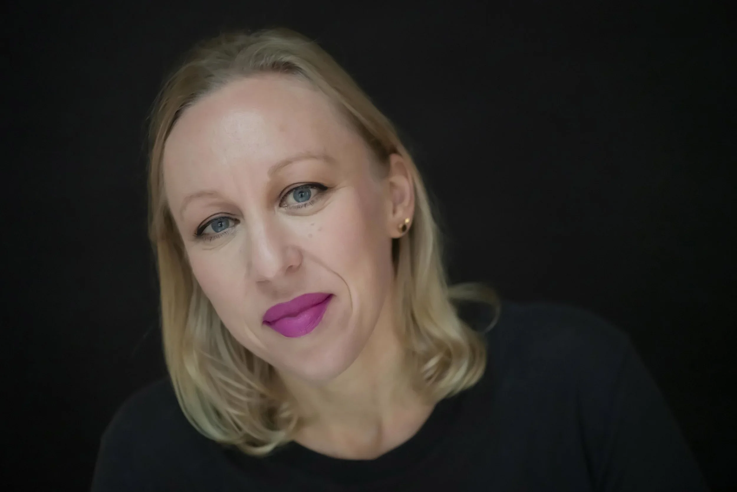 Close-up of a woman with blonde hair, blue eyes, pink lipstick, and small gold earrings, wearing a dark top, against a black background.