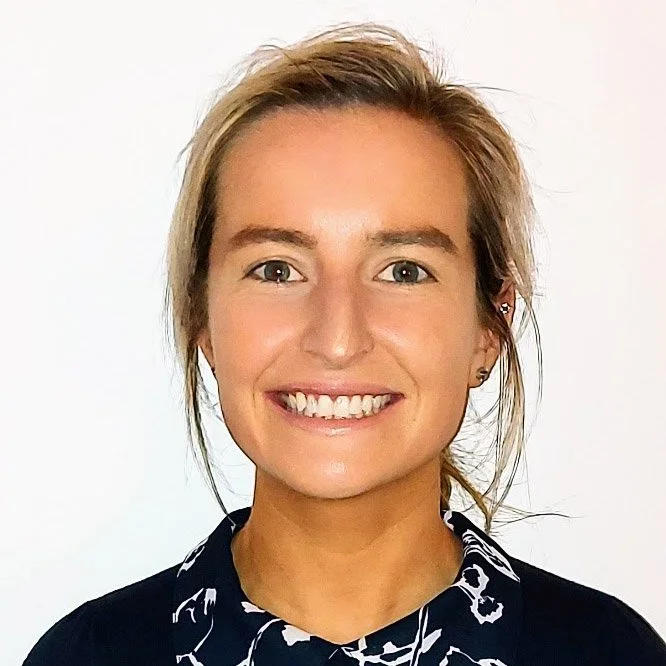 A smiling woman with light skin and blonde hair, wearing a dark top with a white floral pattern, against a plain light background.