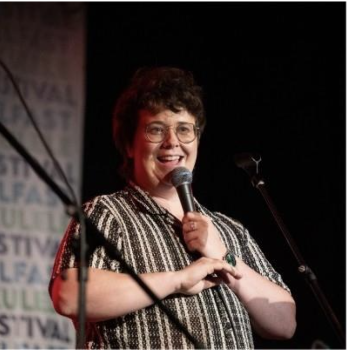 A woman with short curly hair and glasses speaking into a microphone on stage, smiling, wearing a black and white striped shirt.