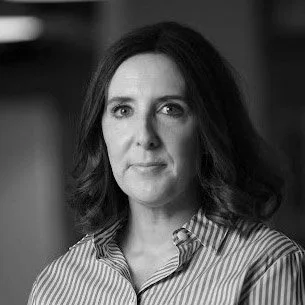 A black and white portrait of a woman with shoulder-length dark hair, wearing a striped collared shirt, looking directly at the camera