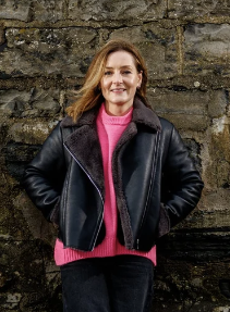 A woman standing outdoors in front of a stone wall, smiling, wearing a black leather jacket over a pink sweater.