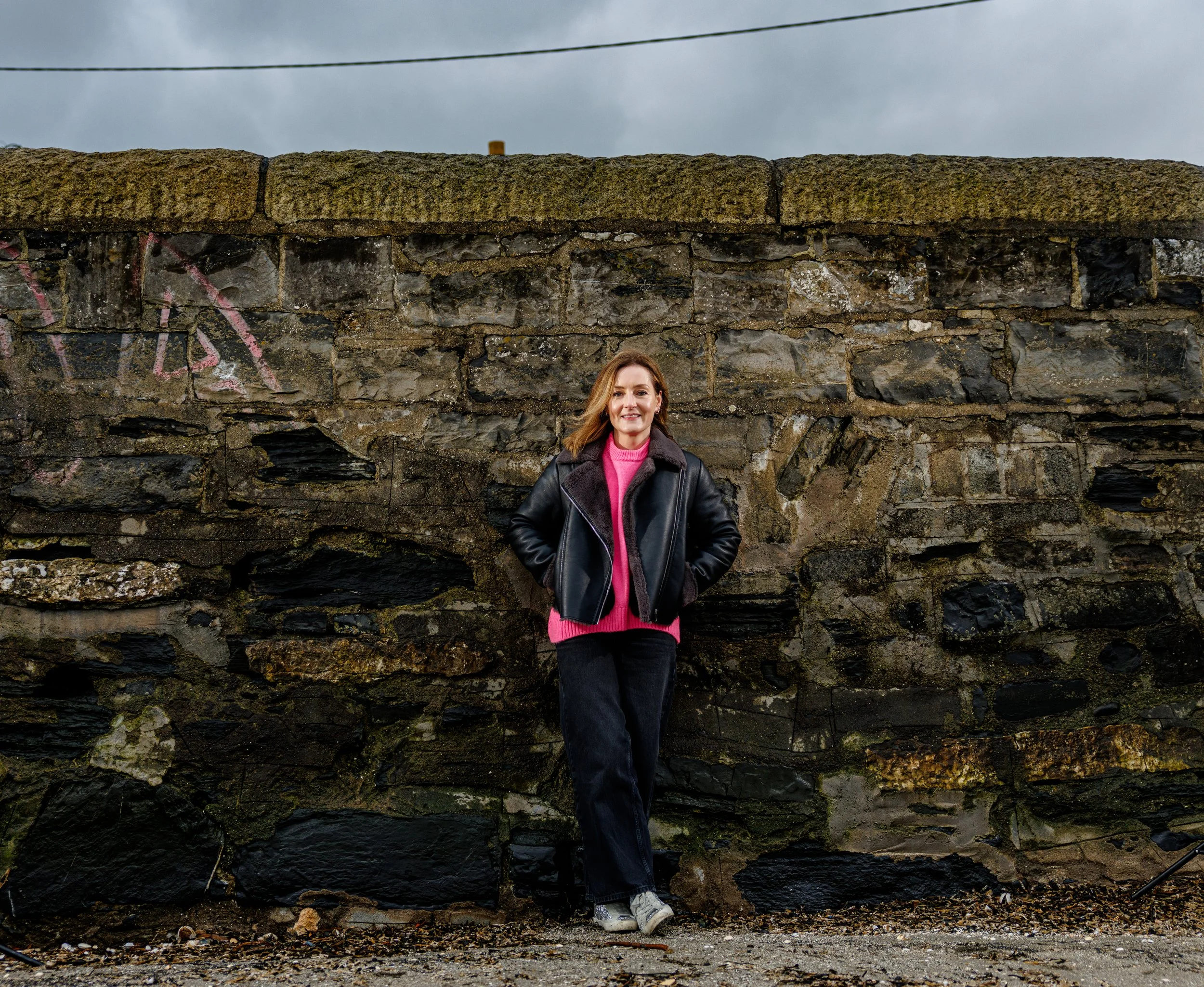 A woman standing outdoors against an old stone wall with graffiti, wearing a black leather jacket, pink sweater, black pants, and white sneakers, smiling at the camera.