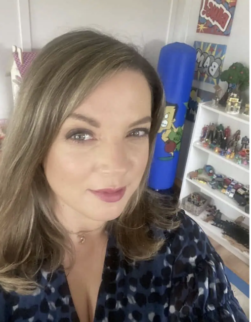A woman taking a selfie in a room with a white shelving unit filled with toys and collectibles, a blue punching bag, and colorful posters on the wall.