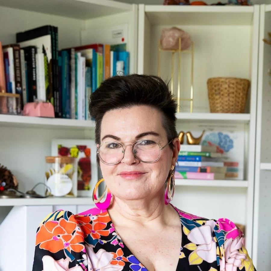 A woman with short dark hair, glasses, and pink earrings, wearing a colorful floral top, smiling in front of a white bookshelf filled with books and decorative items.