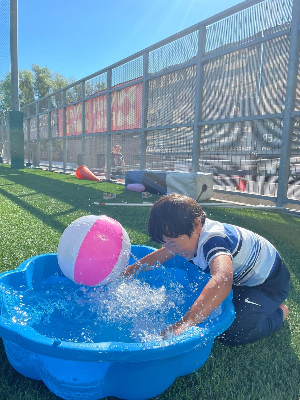A young boy playing with a beach ball in a small blue plastic pool on a grassy field during a sunny day, with a fence and advertisements in the background.