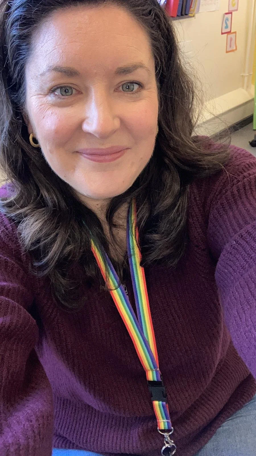 A woman with wavy dark hair, wearing a purple sweater and a rainbow-colored lanyard, smiling at the camera in a classroom or office setting.