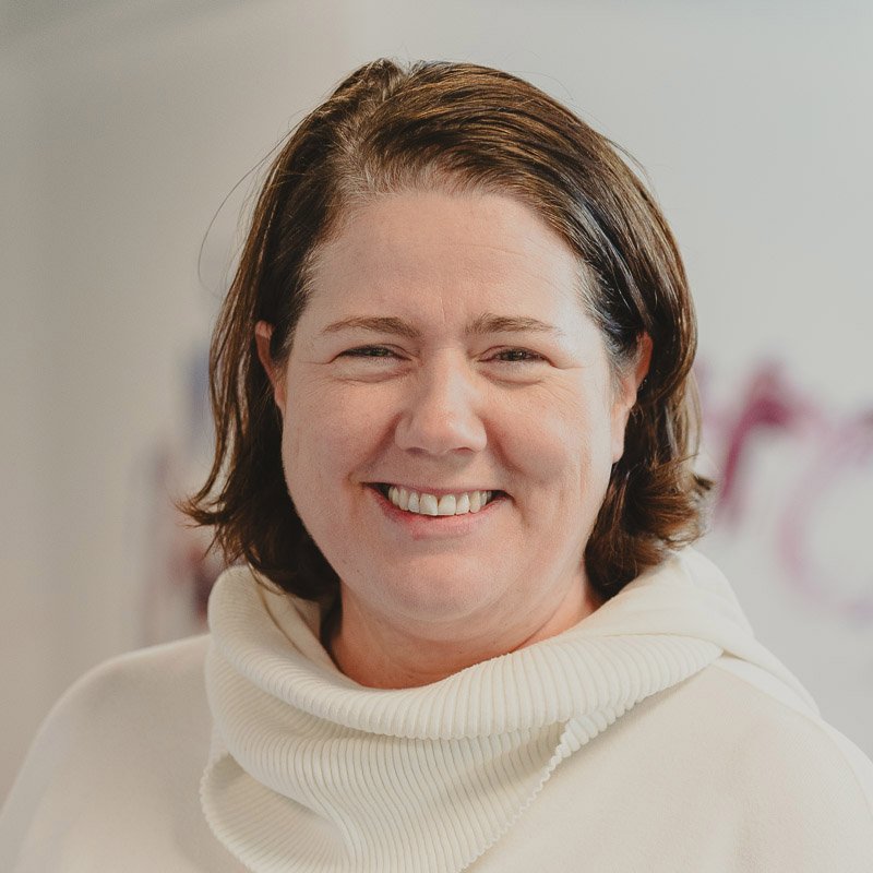 A smiling woman with short brown hair wearing a white turtleneck sweater indoors.