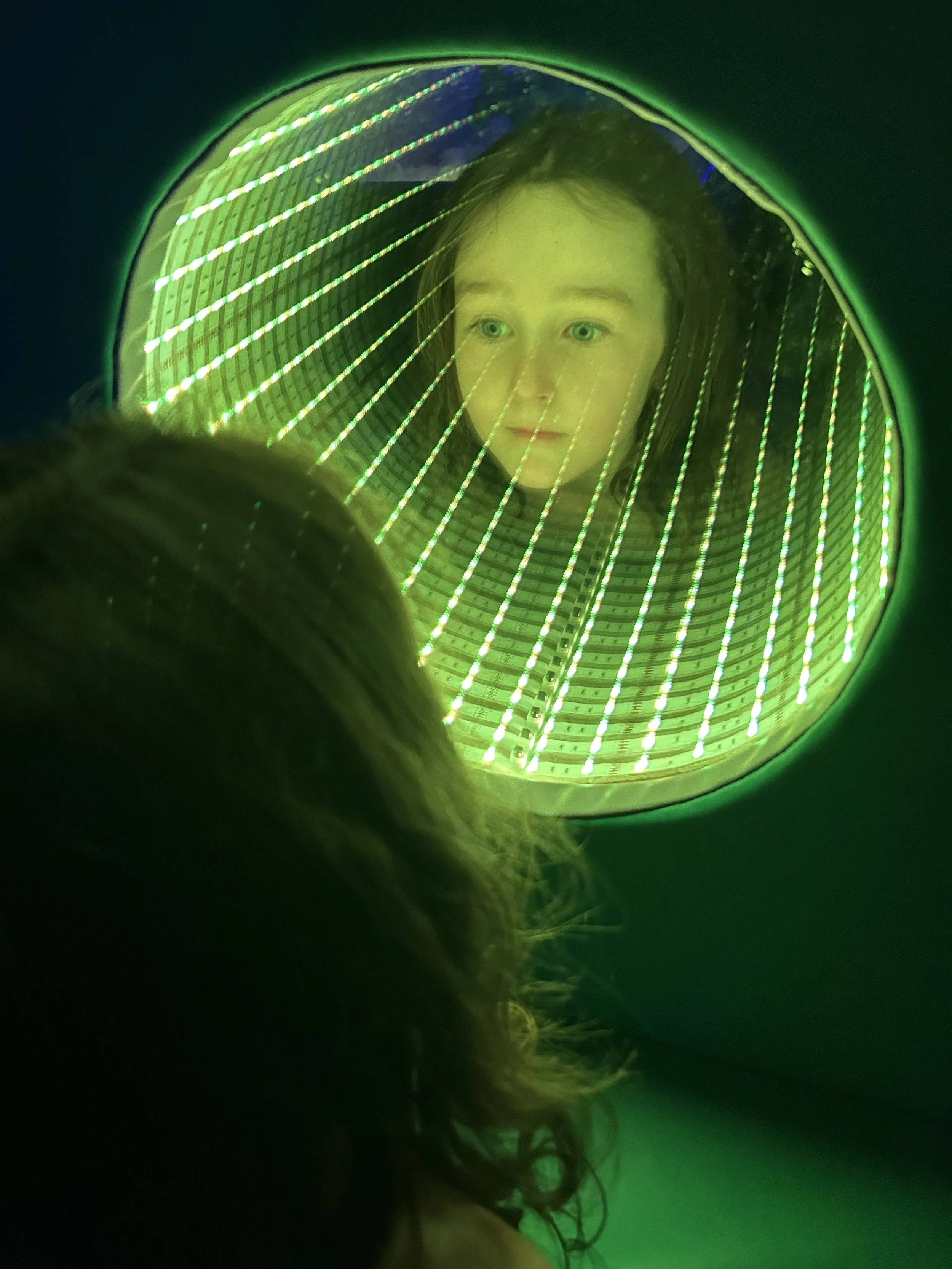 A young girl looking at herself in a mirror with green LED light surrounds, with another person's head in the foreground.