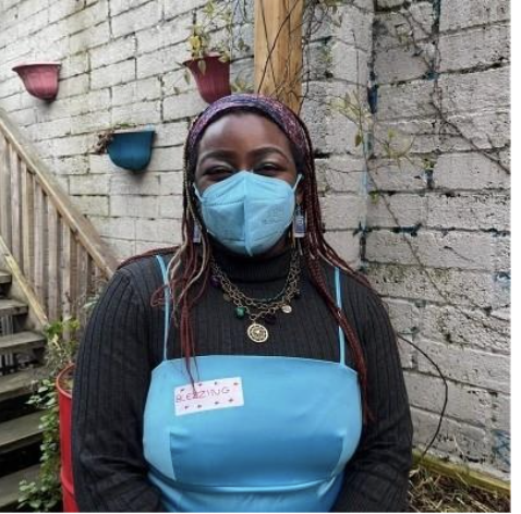 A woman wearing a blue apron, face mask, with jewelry, standing outdoors against a brick wall with colorful plant pots, smiling behind her mask.