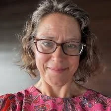 A woman with curly gray hair wearing glasses and a pink patterned top, smiling at the camera.