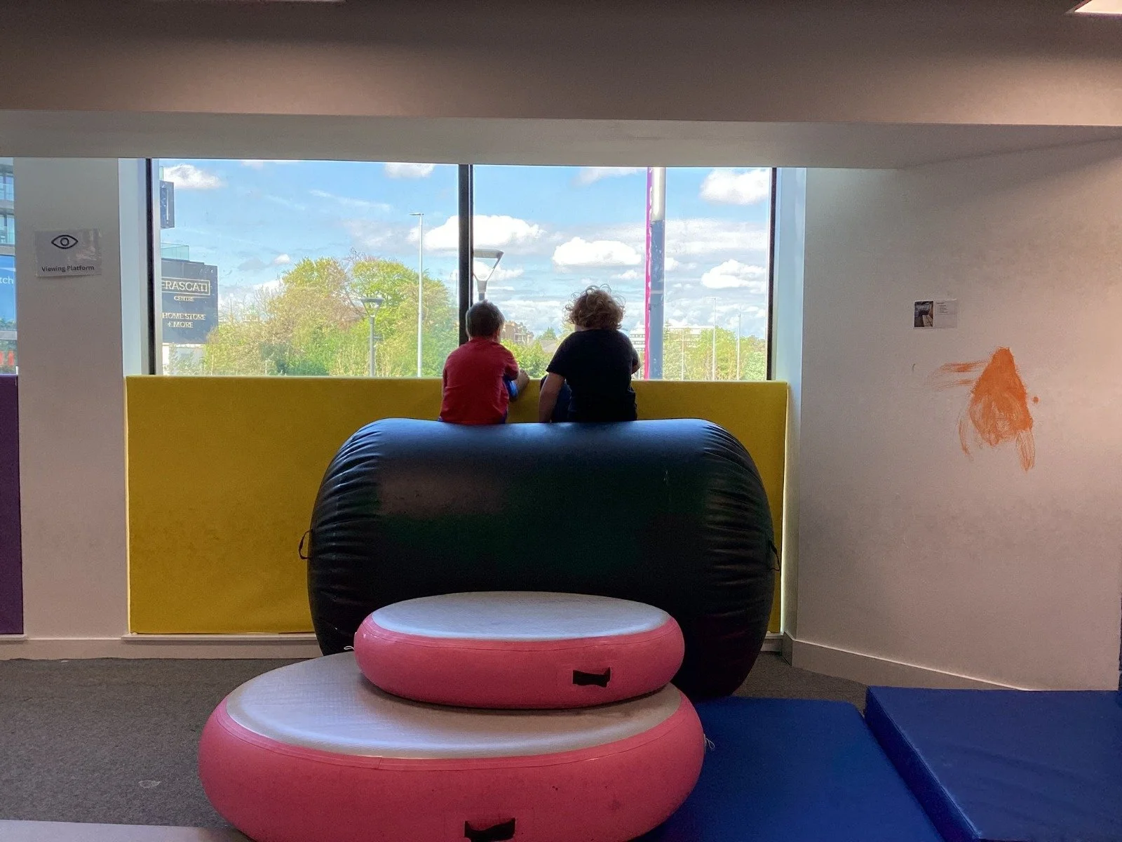 Two children sitting on a yellow bench gazing outside through large window with trees and a cloudy sky, inside a colorful play area with pink and black inflatable structures.