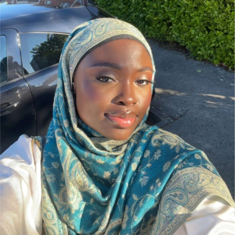 A young African American woman wearing a blue and gold patterned headscarf and a white top, taking a selfie outdoors in sunlight, with a black car and green bushes in the background.